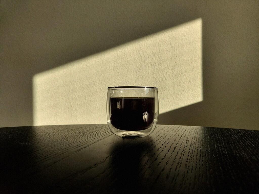 Doppelwandige Glastasse mit frisch gebrühtem Costa Rica Kaffee von Café Granados auf einem dunklen Holztisch; warmes, schräges Sonnenlicht wirft geometrische Schatten an die Wand.